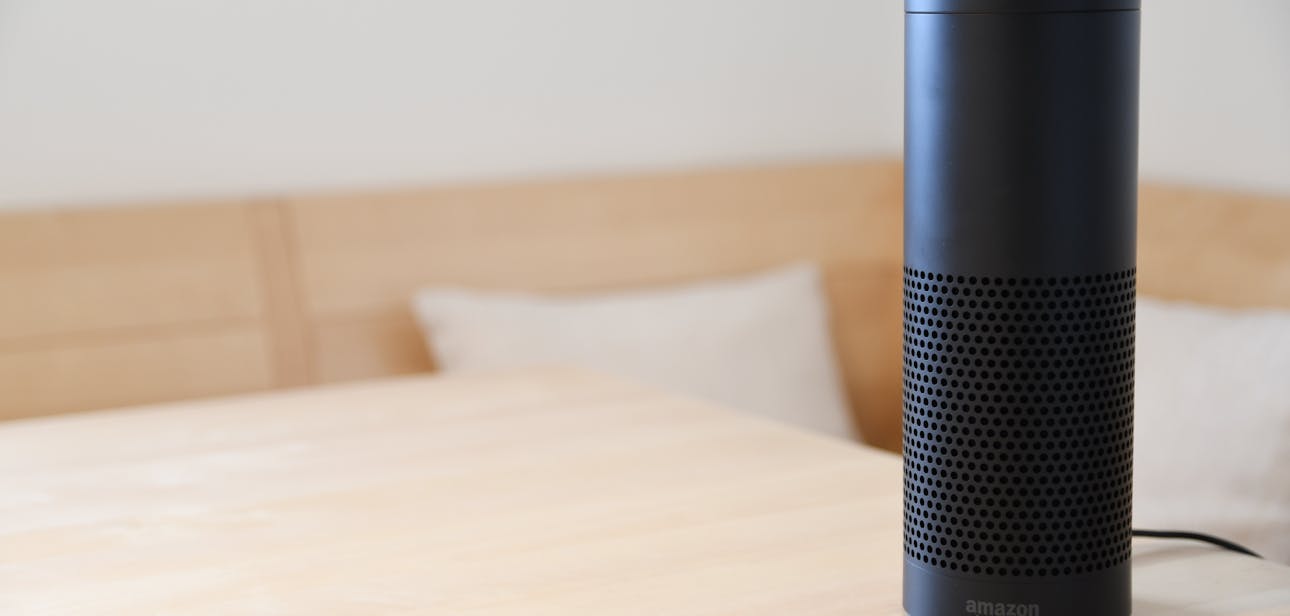 A black smart speaker resting on a light-colored wooden table in a cozy indoor setting.