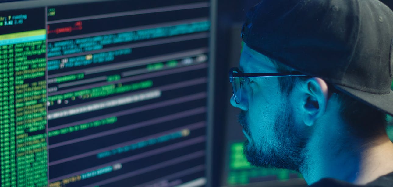 A cybersecurity expert inspecting lines of code on multiple monitors in a dimly lit office.