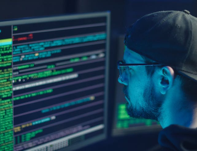 A cybersecurity expert inspecting lines of code on multiple monitors in a dimly lit office.