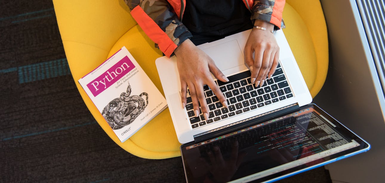 A developer typing code on a laptop with a Python book beside in an office.