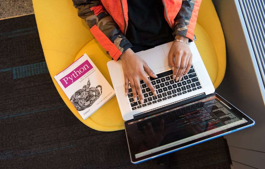 A developer typing code on a laptop with a Python book beside in an office.