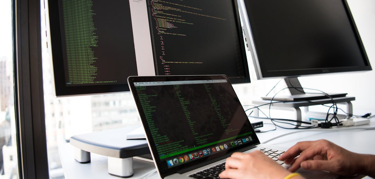 A developer writes code on a laptop in front of multiple monitors in an office setting.