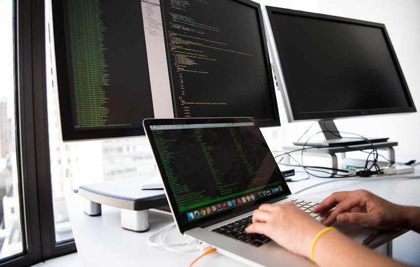 A developer writes code on a laptop in front of multiple monitors in an office setting.