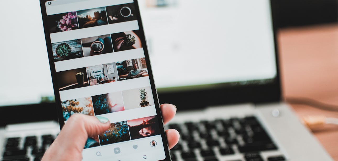 A person uses a smartphone to browse social media in front of a laptop, emphasizing modern technology and lifestyle.