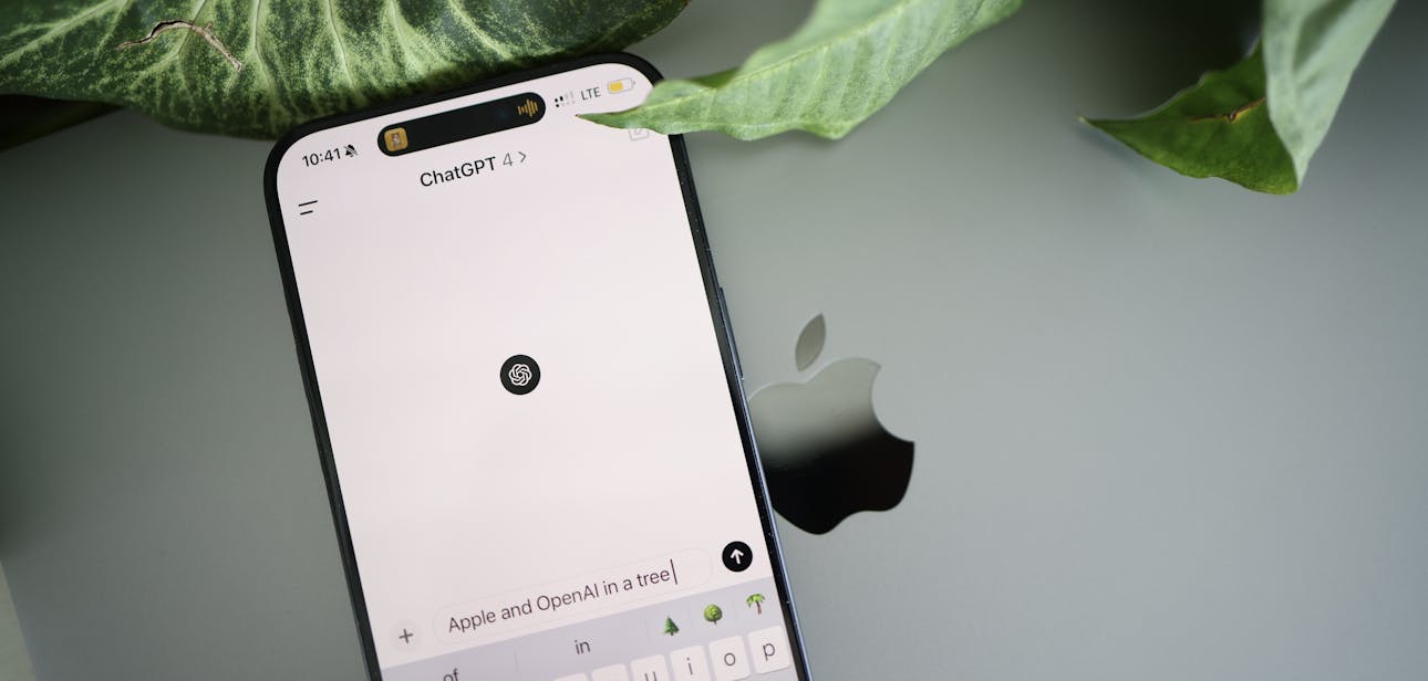 A smartphone shows a ChatGPT interface placed on an Apple laptop in a leafy environment.