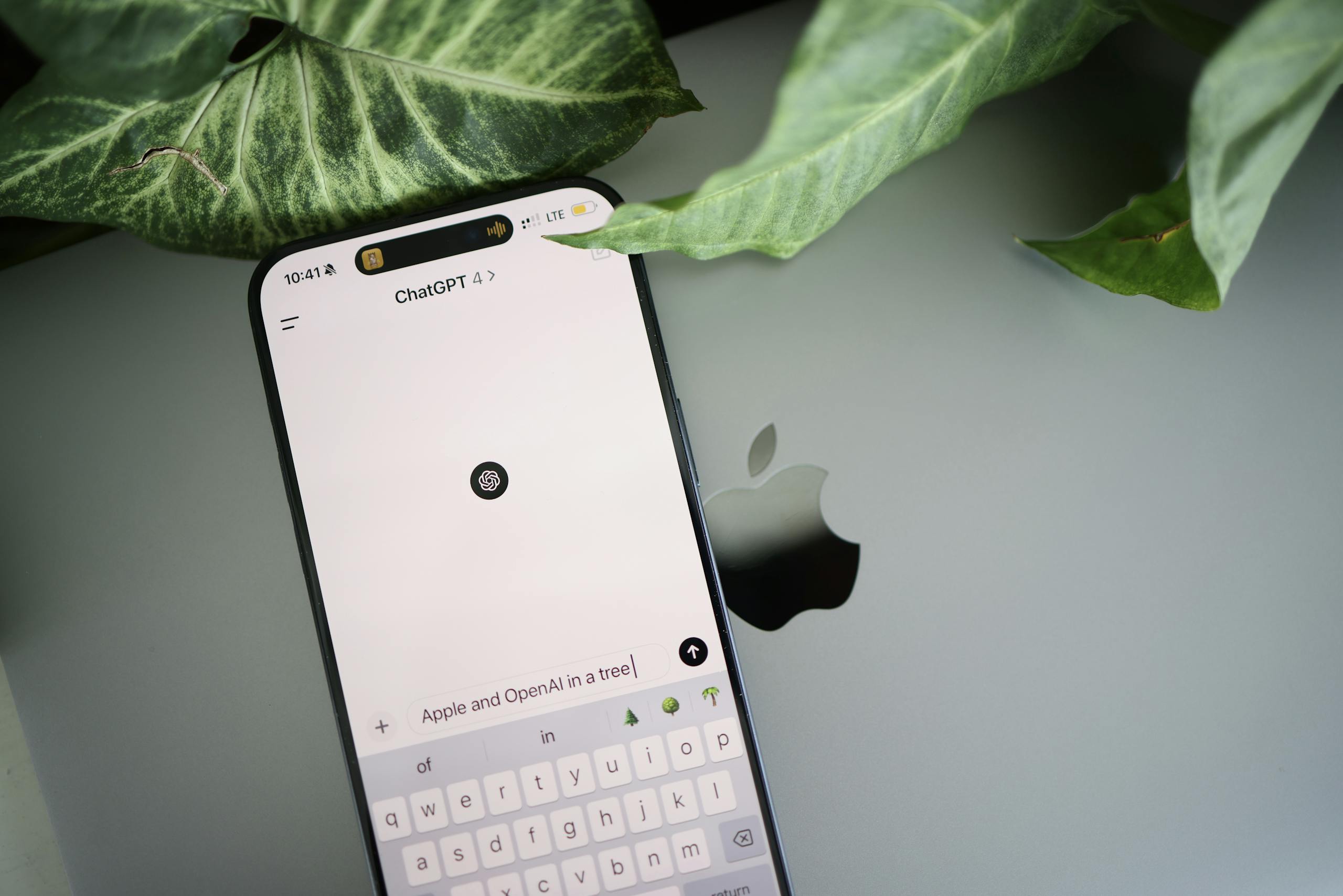 A smartphone shows a ChatGPT interface placed on an Apple laptop in a leafy environment.