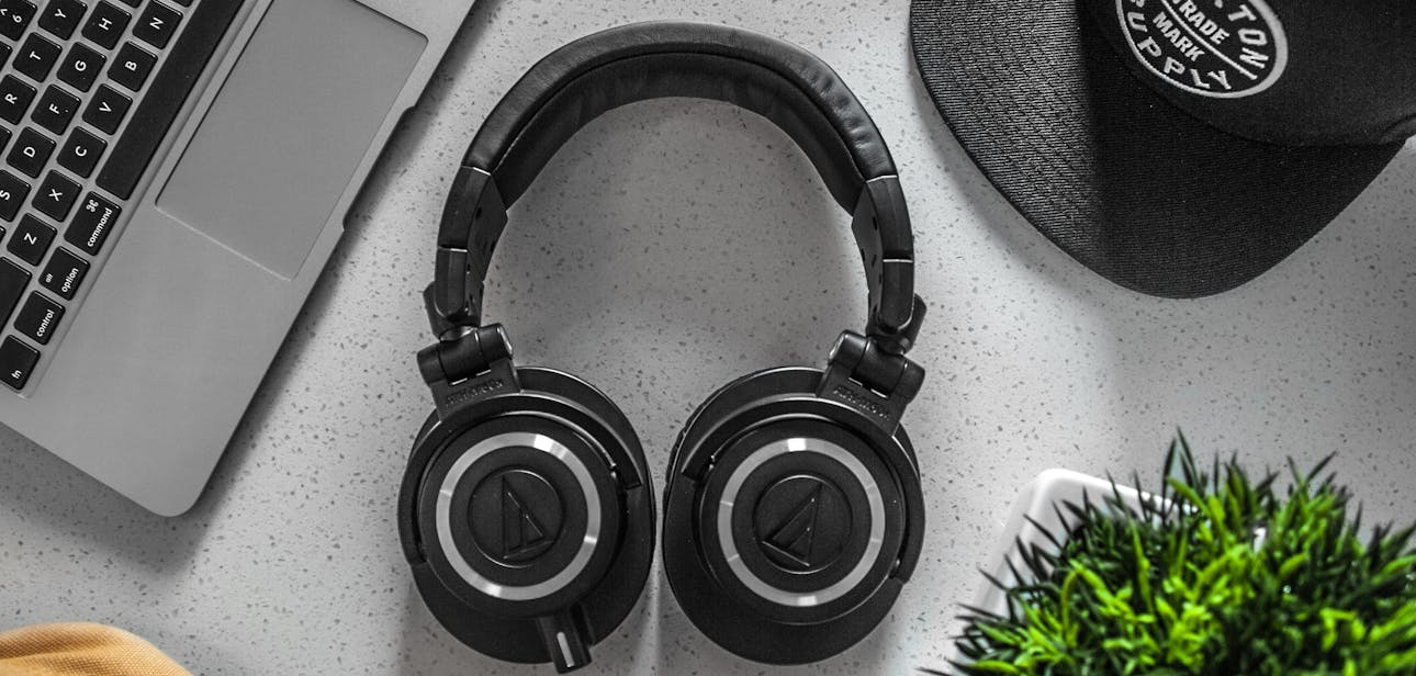 A stylish overhead shot of headphones with a laptop, cap, and decor on a desk.