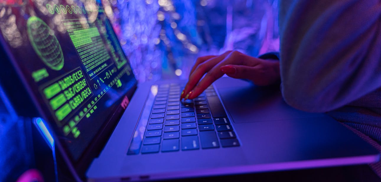Close-up of hands typing on a laptop displaying cybersecurity graphics, illuminated by purple light.