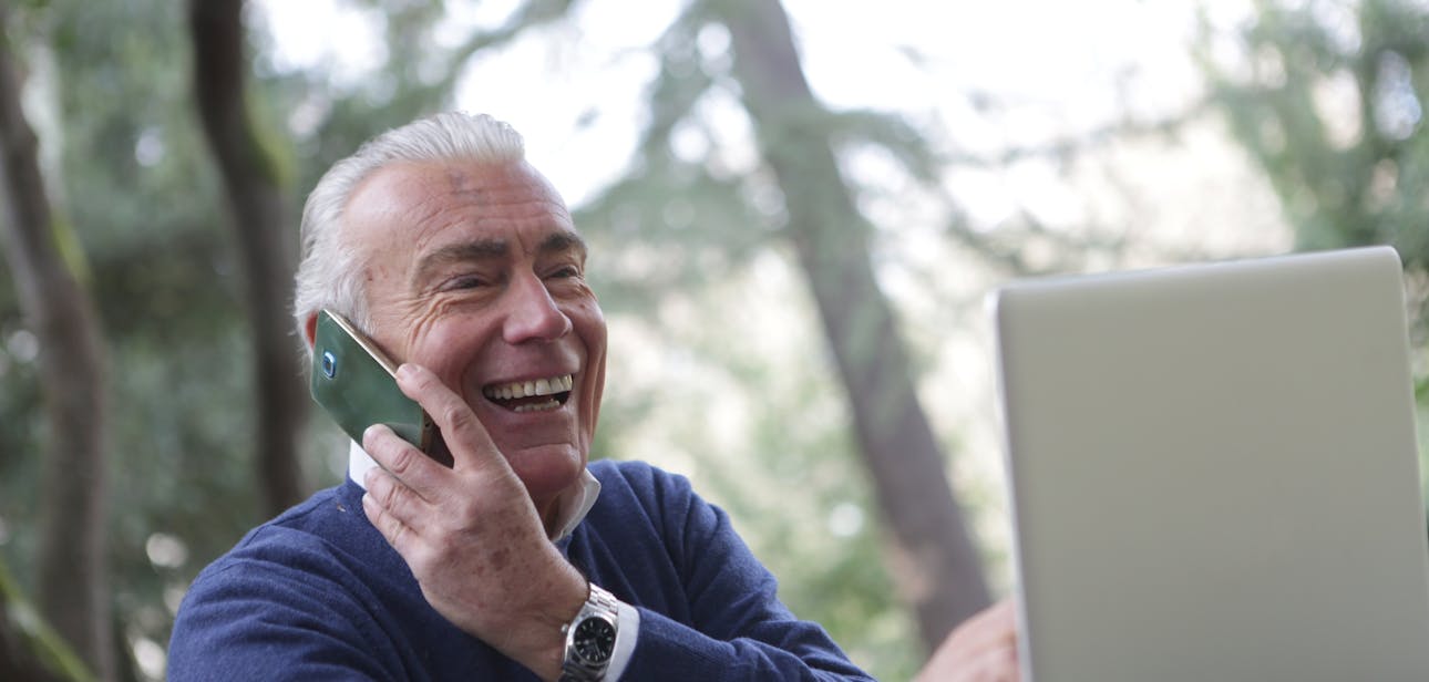 Happy senior man talking on phone and using laptop outdoors.