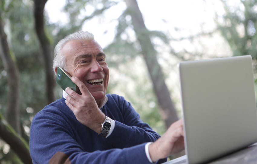 Happy senior man talking on phone and using laptop outdoors.