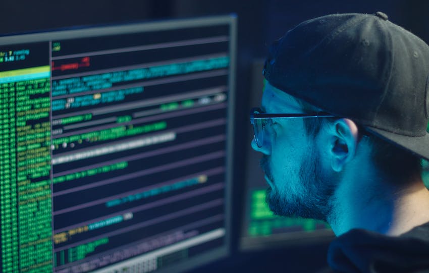 A cybersecurity expert inspecting lines of code on multiple monitors in a dimly lit office.