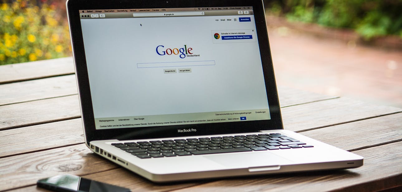 A MacBook Pro displaying Google Search on a wooden table outdoors, next to a smartphone.