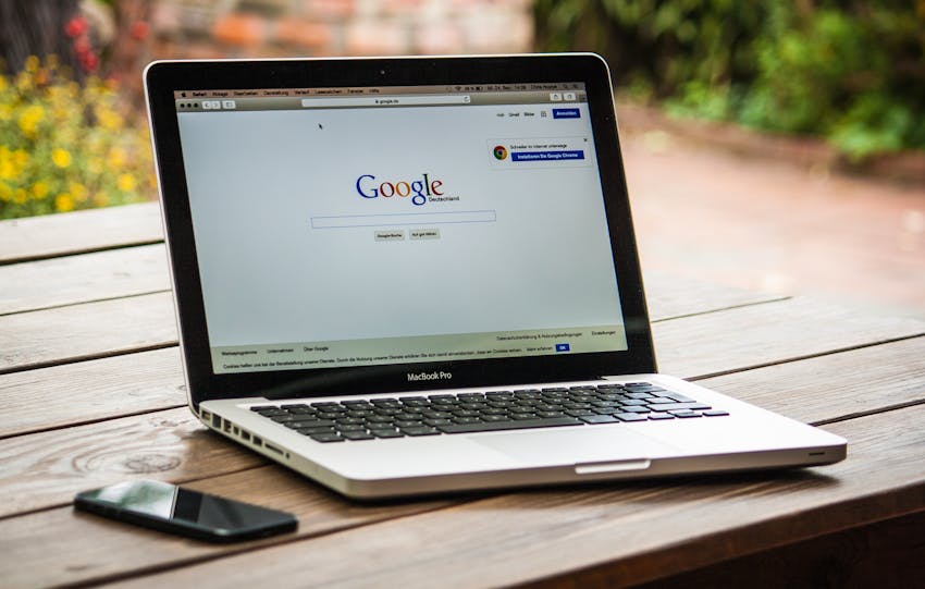 A MacBook Pro displaying Google Search on a wooden table outdoors, next to a smartphone.