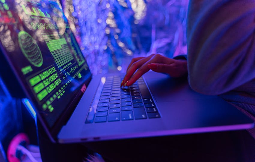 Close-up of hands typing on a laptop displaying cybersecurity graphics, illuminated by purple light.